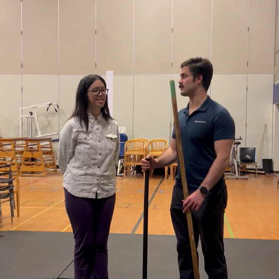 Two people are standing side-by-side in a gym. The person the left has their hands behind their back. The person on the right is leisurely holding a bar in each hand, upright.