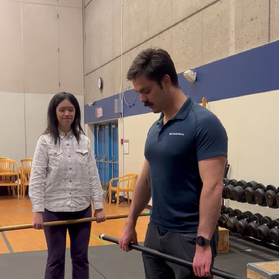 Two people standing in a gym. Both of them are holding a bar with both hands.